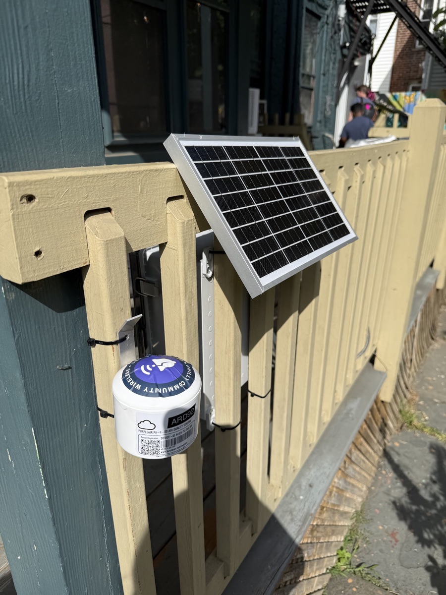 The solar panel on an angled mount above the fence, with the PurpleAir monitor attached to a post below it